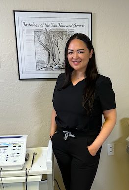 Christy Try in black scrubs standing next to an anatomical chart of skin and hair